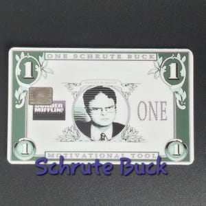 May include: A novelty item resembling a one-dollar bill, featuring a portrait of Dwight Schrute. The design includes the text "ONE SCHRUTE BUCK" and "MOTIVATIONAL TOOL". The item is green and white.