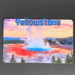 May include: Rectangular magnet featuring a vibrant landscape of Yellowstone National Park. The image showcases a geyser with a blue pool, surrounded by red and orange terrain, under a colorful sky. The word "Yellowstone" is written across the top.
