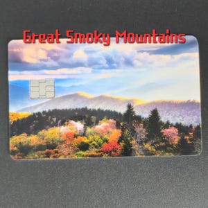 May include: A rectangular card featuring a scenic view of the Great Smoky Mountains. The image showcases a vibrant autumn landscape with colorful trees and rolling hills under a cloudy sky. The words "Great Smoky Mountains" are printed in red at the top.