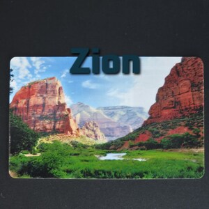 May include: A rectangular magnet featuring a scenic landscape of Zion National Park. The image shows red rock formations, green vegetation, and a blue sky with the word "Zion" in blue letters at the top.
