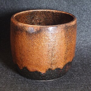 Puede incluir: Un vaso de cerámica hecho a mano con un esmalte de cobre metálico texturizado. La parte inferior de la taza hace la transición a un acabado negro moteado. La taza cilíndrica tiene una abertura ancha y un diseño simple y moderno.