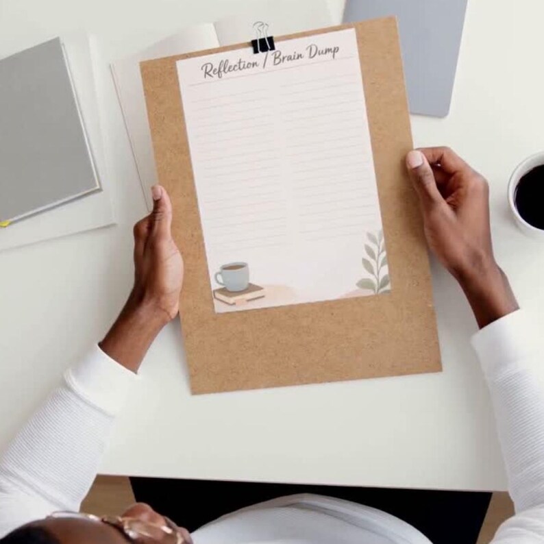 May include: A brown clipboard holding a white sheet of paper with the text Reflection / Brain Dump at the top. The paper features horizontal lines, a small illustration of a light blue mug on a book, and a green leafy plant graphic in the corner.