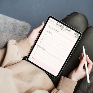 May include: A digital tablet displaying a Weekly Goals planner template with sections for Top Goals, Action Steps, and Notes. The interface features a soft beige background, dark text, and a small green leaf illustration on the right side.