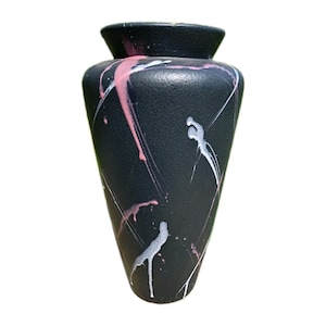 Puede incluir: Un jarrón negro mate con forma cónica y estrecha, que presenta un borde ancho y acampanado. La superficie tiene un acabado texturizado decorado con salpicaduras de pintura abstractas e irregulares en tonos rosa suave y blanco brillante.