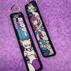May include: Two rectangular keychains with metal rings. One features the Japanese text "Mewtwo" in teal and white, with a white and purple character illustration. The other keychain displays a white and purple character illustration on a navy background.