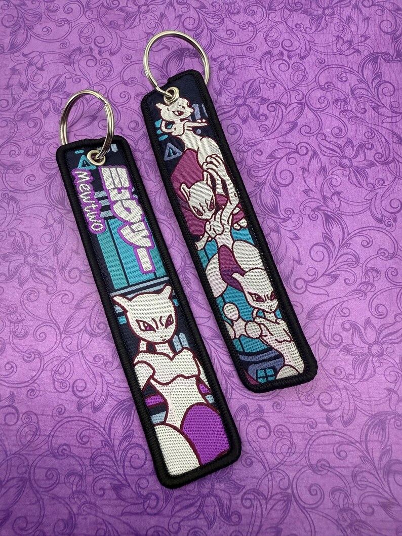 May include: Two rectangular keychains with metal rings. One features the Japanese text "Mewtwo" in teal and white, with a white and purple character illustration. The other keychain displays a white and purple character illustration on a navy background.