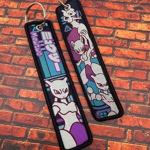 May include: Two embroidered keychains featuring a purple and white Mewtwo design. One keychain has the text "Mewtwo" in a teal and purple design. The other keychain has a Mewtwo graphic with a purple background.
