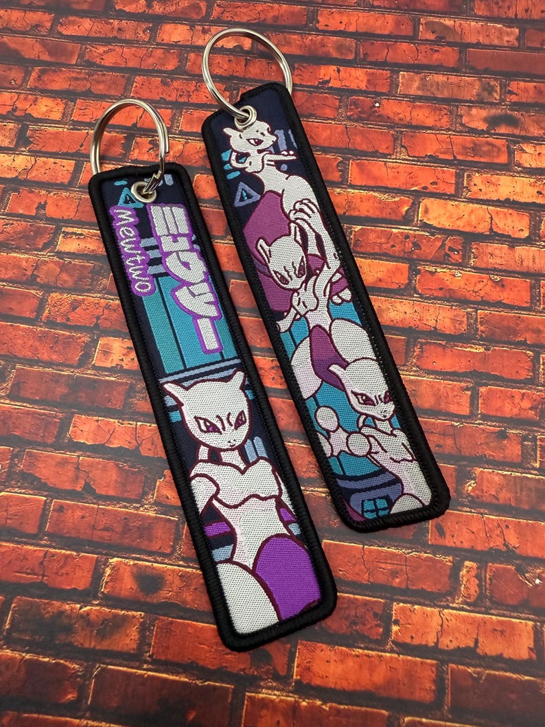 May include: Two embroidered keychains featuring a purple and white Mewtwo design. One keychain has the text "Mewtwo" in a teal and purple design. The other keychain has a Mewtwo graphic with a purple background.