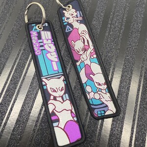 May include: Two rectangular keychains with a black border and silver keyring. One keychain features the text "Mewtwo" in pink and Japanese characters over a teal and blue background. The other keychain has a Mewtwo graphic in white, pink, and purple.