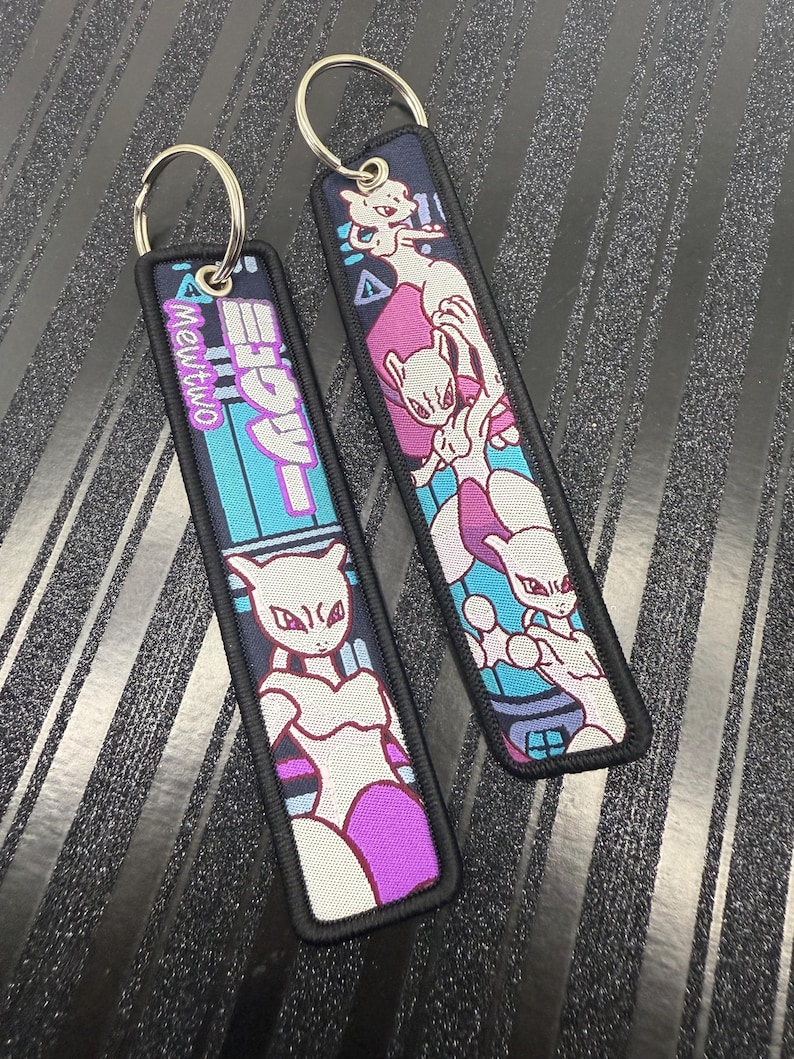 May include: Two rectangular keychains with a black border and silver keyring. One keychain features the text "Mewtwo" in pink and Japanese characters over a teal and blue background. The other keychain has a Mewtwo graphic in white, pink, and purple.