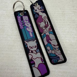 May include: Two rectangular keychains with silver key rings. The keychains feature a purple, gray, and teal design with the text "Mewtwo" in Japanese and an image of the character. The keychains have a black border.