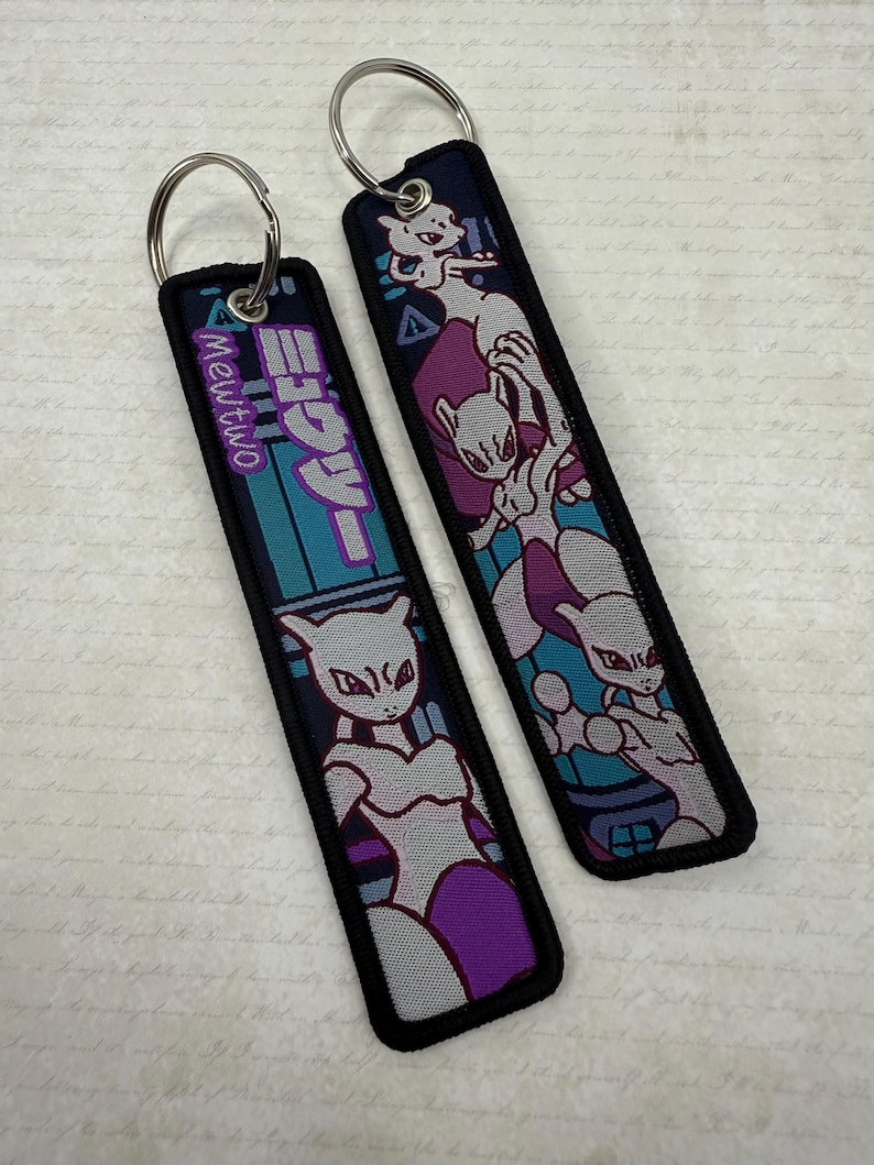 May include: Two rectangular keychains with silver key rings. The keychains feature a purple, gray, and teal design with the text "Mewtwo" in Japanese and an image of the character. The keychains have a black border.
