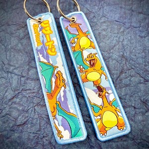 May include: Two rectangular keychains with a light blue border and a metal ring. Each keychain features a cartoon Charizard design in orange, yellow, and teal, set against a light blue and white background. The text "Charizard" is visible on one keychain.