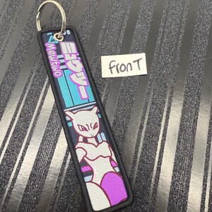May include: A rectangular keychain with a metal ring. The keychain features a cartoon image of Mewtwo in white, purple, and black. Japanese text and the word "Mewtwo" are also visible on the keychain.
