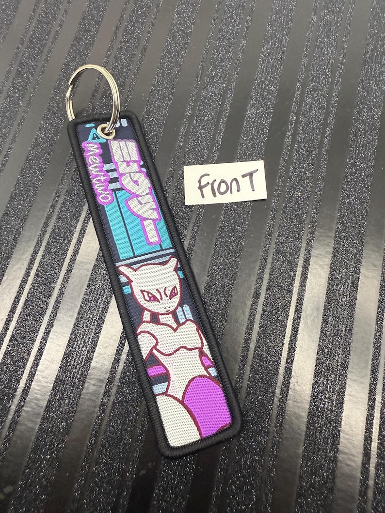 May include: A rectangular keychain with a metal ring. The keychain features a cartoon image of Mewtwo in white, purple, and black. Japanese text and the word "Mewtwo" are also visible on the keychain.