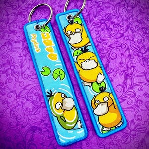 May include: Two rectangular keychains with a blue border and a silver key ring. The keychains feature a yellow cartoon duck character with the text "Psyduck" in yellow and Japanese characters. The background is a purple floral pattern.