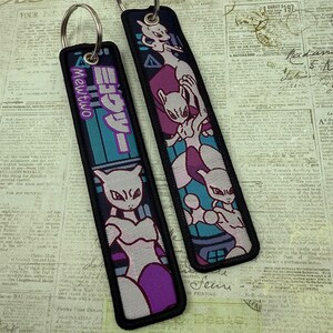 May include: Two rectangular keychains with metal rings. One features the text "Mewtwo" and Japanese characters, with a purple and teal design. The other has a Mewtwo graphic in purple, pink, and teal. Both have black borders.