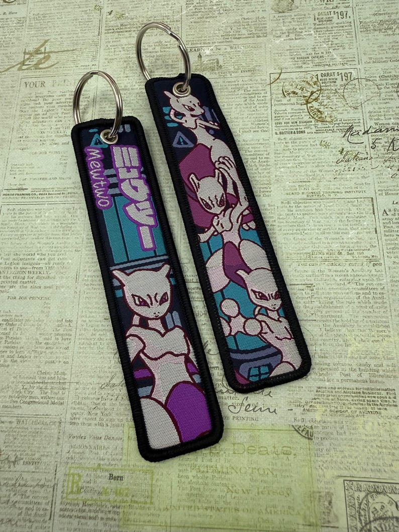May include: Two rectangular keychains with metal rings. One features the text "Mewtwo" and Japanese characters, with a purple and teal design. The other has a Mewtwo graphic in purple, pink, and teal. Both have black borders.