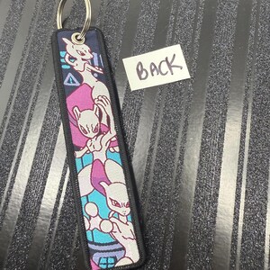 May include: A rectangular keychain with a metal ring. The keychain features a colorful design of a cartoon character in shades of white, purple, and blue. The edges are black, and the word "BACK" is written on a white label.