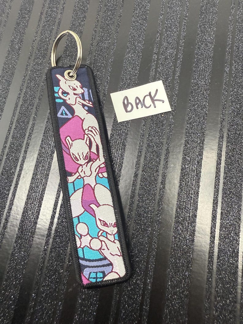 May include: A rectangular keychain with a metal ring. The keychain features a colorful design of a cartoon character in shades of white, purple, and blue. The edges are black, and the word "BACK" is written on a white label.