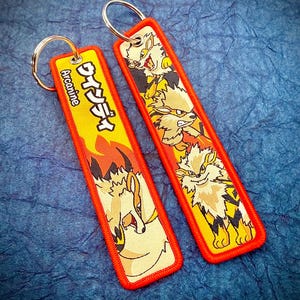 May include: Two embroidered keychains with a red border and a yellow background. One keychain has Japanese text and the word "Arcanine". The other keychain has a graphic of a cartoon dog. Both keychains have a silver key ring.