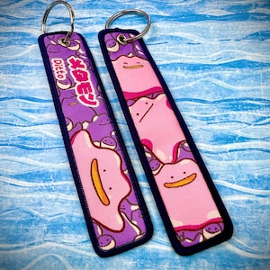 May include: Two rectangular keychains featuring the Pokémon Ditto. One keychain has the word "Ditto" and Japanese text. The other keychain has a pink Ditto design. Both keychains have a purple background and a silver key ring.