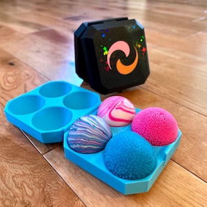 Puede incluir: Una bandeja cuadrada de color azul brillante con cuatro hendiduras circulares que contienen cuatro esferas distintas: una jaspeada rosa y blanca, una jaspeada multicolor, una texturizada rosa brillante y una texturizada azul brillante. Detrás hay una caja negra.