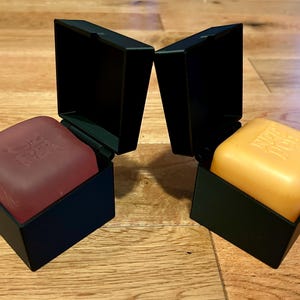 Puede incluir: Dos pastillas de jabón cuadradas, una de color rojo oscuro y otra amarilla, cada una en una caja negra abierta con bisagras. Las pastillas tienen texto en relieve en la parte superior. Los recipientes son negro mate con base cuadrada.