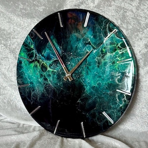 Puede incluir: Reloj de pared redondo con un bisel negro y un diseño abstracto fascinante en tonos verdes y turquesas. La esfera del reloj presenta marcadores de hora y manecillas plateadas, creando una estética moderna. Un elemento decorativo para cualquier habitación.