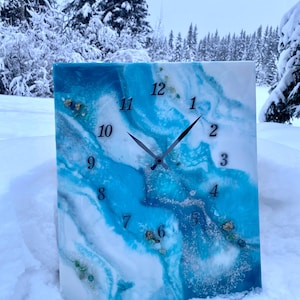 May include: A square wall clock with a blue and white resin design. The clock face features black hands and black numerals. The design includes swirls of blue and white with embedded decorative elements. The clock is set against a snowy backdrop.