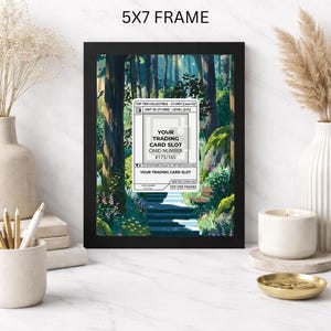 May include: A black 5x7 frame displaying a forest scene with a path and a trading card slot. The card slot has text that reads "YOUR TRADING CARD SLOT CARD NUMBER #175/165". The frame is on a white surface.