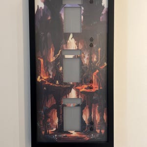 Puede incluir: Una cubierta de interruptor de luz enmarcada en negro con un dise&ntilde;o de paisaje de fantas&iacute;a. La obra de arte presenta una escena cavernosa y ardiente con lava y formaciones rocosas. Tiene tres aberturas para interruptores.