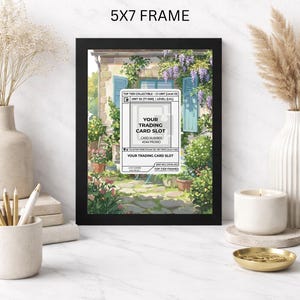 May include: A black 5x7 frame displaying a colorful print of a European-style building with blue shutters and lush greenery. The frame includes a white card slot for trading cards with text that reads "YOUR TRADING CARD SLOT".