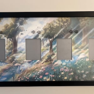 May include: A black rectangular picture frame with four openings. The background art depicts a forest scene with trees, a stream, and flowers. The frame is mounted on a white wall.