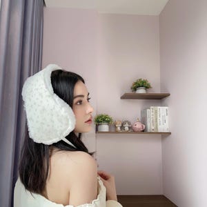 Puede incluir: Orejeras blancas con un estampado floral. Las orejeras están hechas de un material suave y esponjoso y están diseñadas para cubrir las orejas. Estas orejeras son un accesorio de invierno, que proporciona calidez y estilo. Fondo de pared rosa claro.