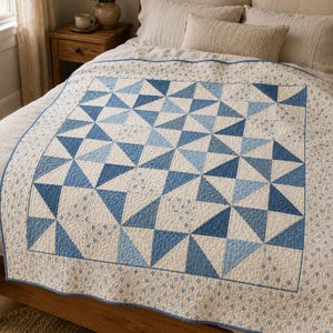 Op de afbeelding: Een quilt met een geometrisch patroon van blauwe en witte driehoeken gerangschikt in een centraal vierkant blok. De rand en de buitenste delen zijn gemaakt van crèmekleurige stof met een kleine blauwe bloemenprint, afgewerkt met een blauwe bies.