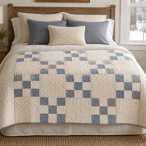 May include: A quilt featuring a repeating pattern of cream-colored squares and blue nine-patch blocks. The fabric has a visible stitched texture throughout, with a solid blue border trim along the edges, covering the surface of a bed with pillows.