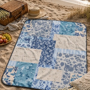 May include: A blue and white patchwork picnic blanket with floral patterns, set on a sandy beach. A wicker picnic basket, straw hat, and fruit basket are nearby, suggesting a relaxing outdoor scene.