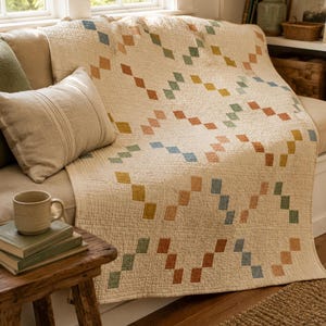 Op de afbeelding: Een cr&egrave;mekleurige gewatteerde plaid met een herhalend geometrisch patroon van kleine, vierkante stiksels in gedempte tinten blauw, groen, geel en terracotta oranje, gerangschikt in een diagonaal, in elkaar grijpend schakelontwerp.