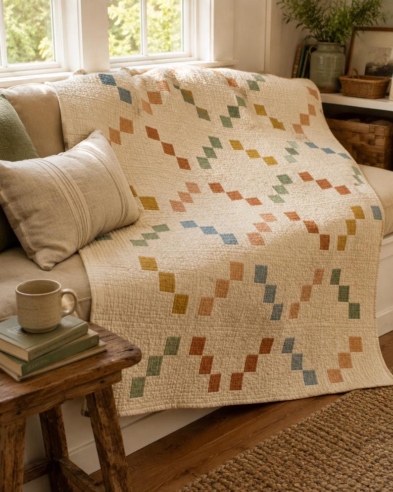 Op de afbeelding: Een cr&egrave;mekleurige gewatteerde plaid met een herhalend geometrisch patroon van kleine, vierkante stiksels in gedempte tinten blauw, groen, geel en terracotta oranje, gerangschikt in een diagonaal, in elkaar grijpend schakelontwerp.