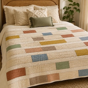 Puede incluir: Una colcha de color crema con un patrón repetitivo de parches rectangulares en tonos apagados de verde salvia, terracota, amarillo mostaza, azul polvoriento y topo. La colcha tiene una textura acolchada visible y cubre una cama con almohadas.