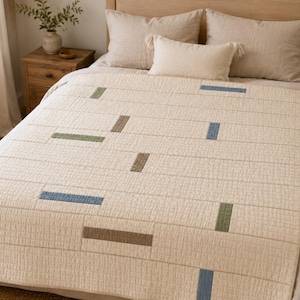 May include: A cream-colored quilted bedspread featuring a grid pattern of horizontal and vertical lines. The quilt is accented with scattered rectangular patches in muted shades of sage green, dusty blue, and earthy brown across the surface.
