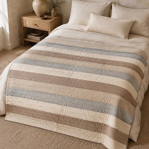 May include: A quilted bedspread featuring horizontal stripes in shades of beige, tan, and muted blue. The bedding set includes two standard pillows and one rectangular lumbar pillow, all in a solid, neutral beige tone, covering a bed.