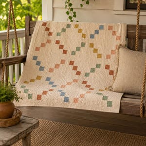 Op de afbeelding: Een cr&egrave;mekleurige quilt met een herhalend geometrisch patroon van kleine, vierkante blokjes in gedempte tinten blauw, groen, geel en terracotta. De stof heeft een zichtbaar geweven textuur en ligt gedrapeerd over een houten schommelbank.