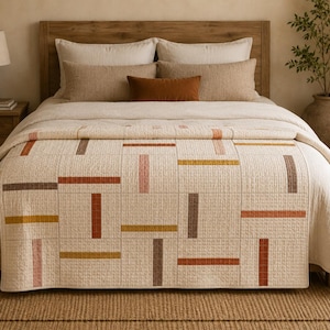 May include: A cream-coloured quilt featuring a grid pattern of stitched squares. The quilt is decorated with horizontal and vertical rectangular blocks in shades of terracotta, mustard yellow, and brown, creating a geometric design across the surface.
