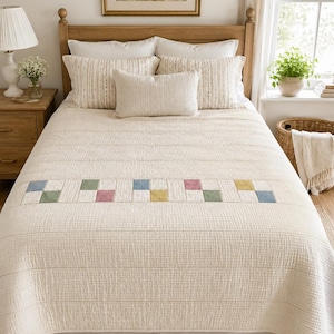 May include: A cream-coloured quilted bedspread featuring a horizontal band of small, multi-coloured square patches in shades of blue, green, pink, and yellow. The bedding set includes two matching rectangular pillows and one smaller central lumbar pillow.