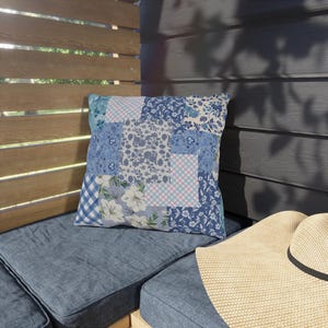 Puede incluir: Cojín decorativo cuadrado con un diseño de patchwork en tonos azules, blancos y rosa claro. El cojín presenta patrones florales y a cuadros. Descansa sobre un cojín azul, con un sombrero de paja en primer plano.