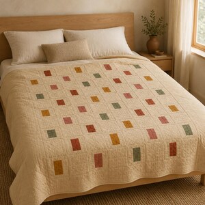 May include: A cream-colored quilt featuring a grid pattern of small, rectangular patches in muted shades of mustard yellow, sage green, and terracotta red. The quilt has a visible stitched texture and covers a bed with two large pillows.