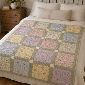 May include: A patchwork quilt featuring a grid of square fabric panels in pastel shades of pink, yellow, blue, and lavender, each decorated with a small floral print. The squares are separated by sage green sashing and small cream cornerstones.