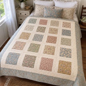 Può includere: Una trapunta patchwork caratterizzata da una griglia di pannelli quadrati in tessuto con vari motivi floreali in tonalità tenui di blu, rosa, verde e giallo, separati da fasce color crema e rifinita con un ampio bordo a motivi floreali azzurri.